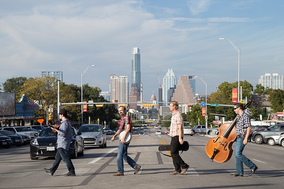 Four Austin musicians carry a banjo, guitar, and upright bass across South Congress with the downtown skyline behind them