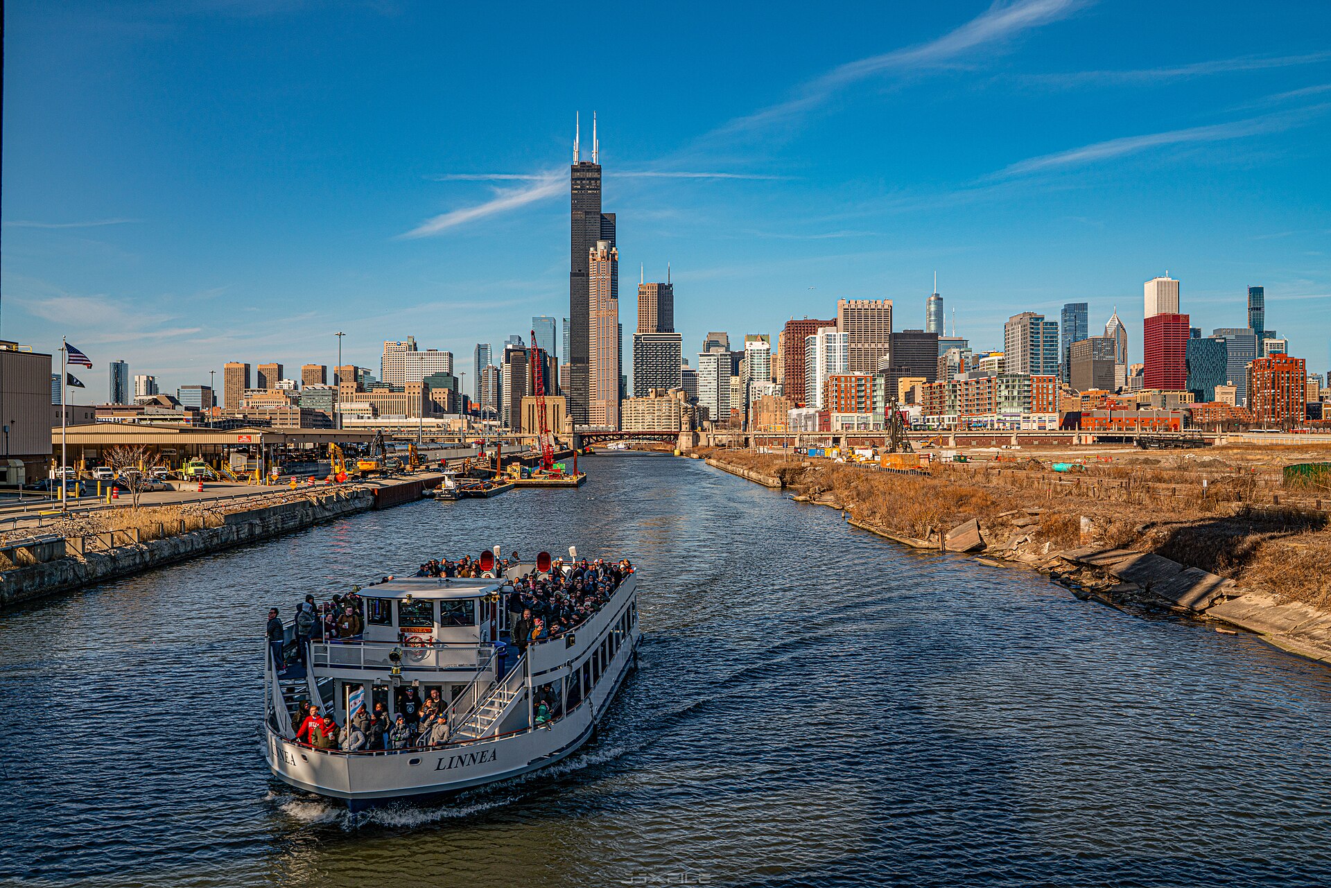 A crowded Chicago River tour boat approaches the skyline with the Willis Tower rising beyond