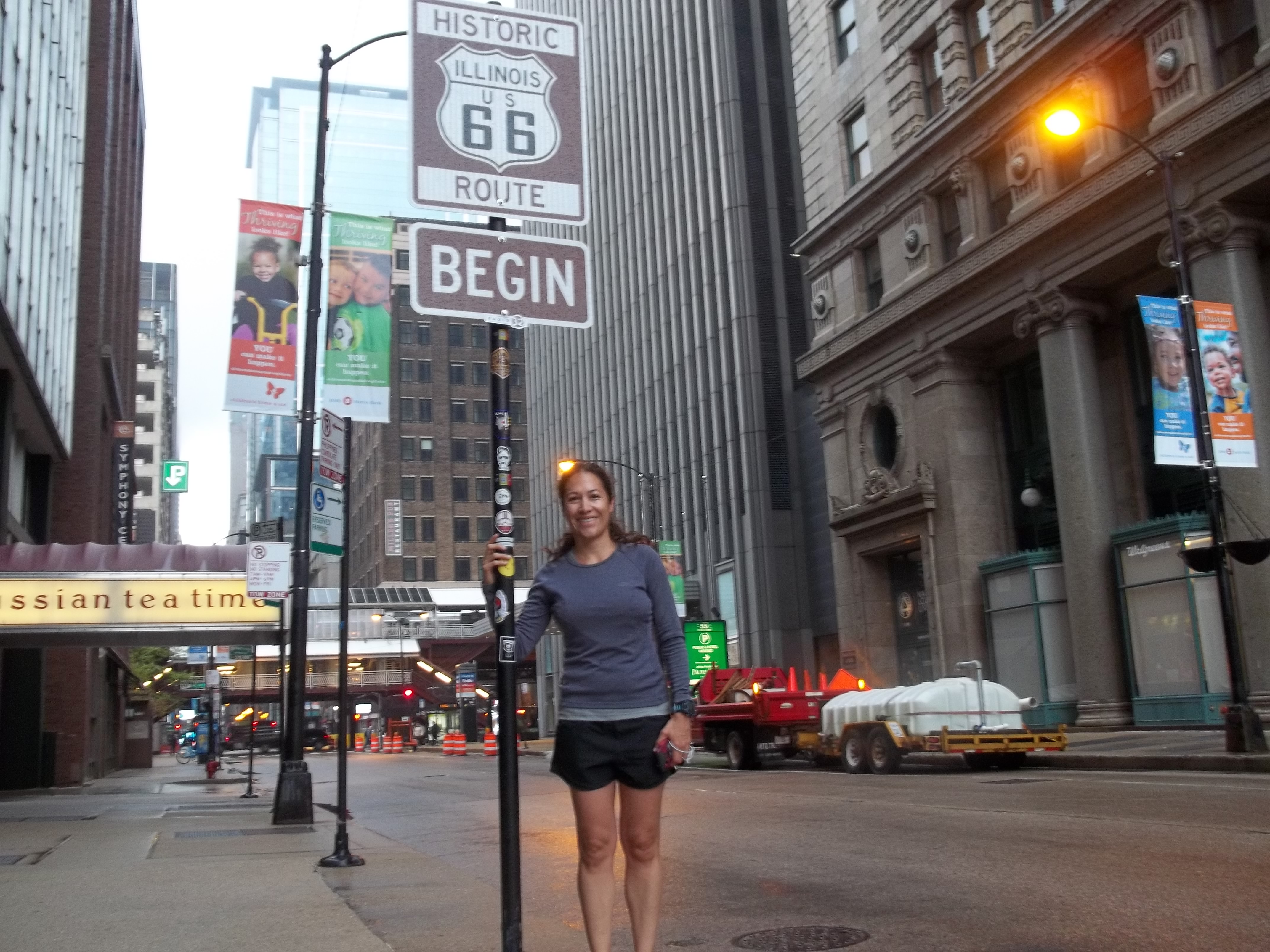 A traveler stands beneath Chicago's Historic Route 66 Begin sign amid downtown architecture