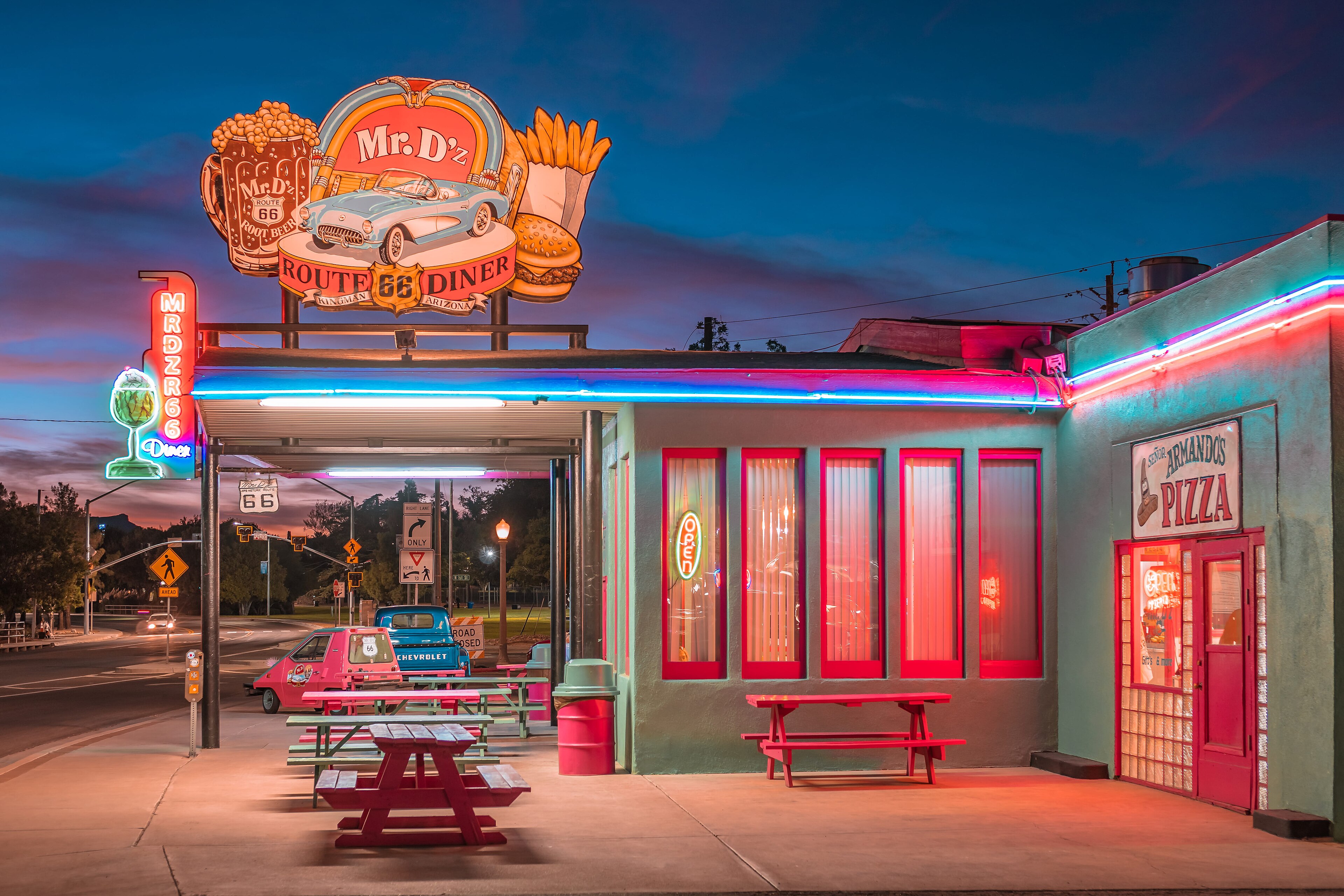 An operating Route 66 diner glows with neon, food signs, outdoor tables, and parked local cars at dusk