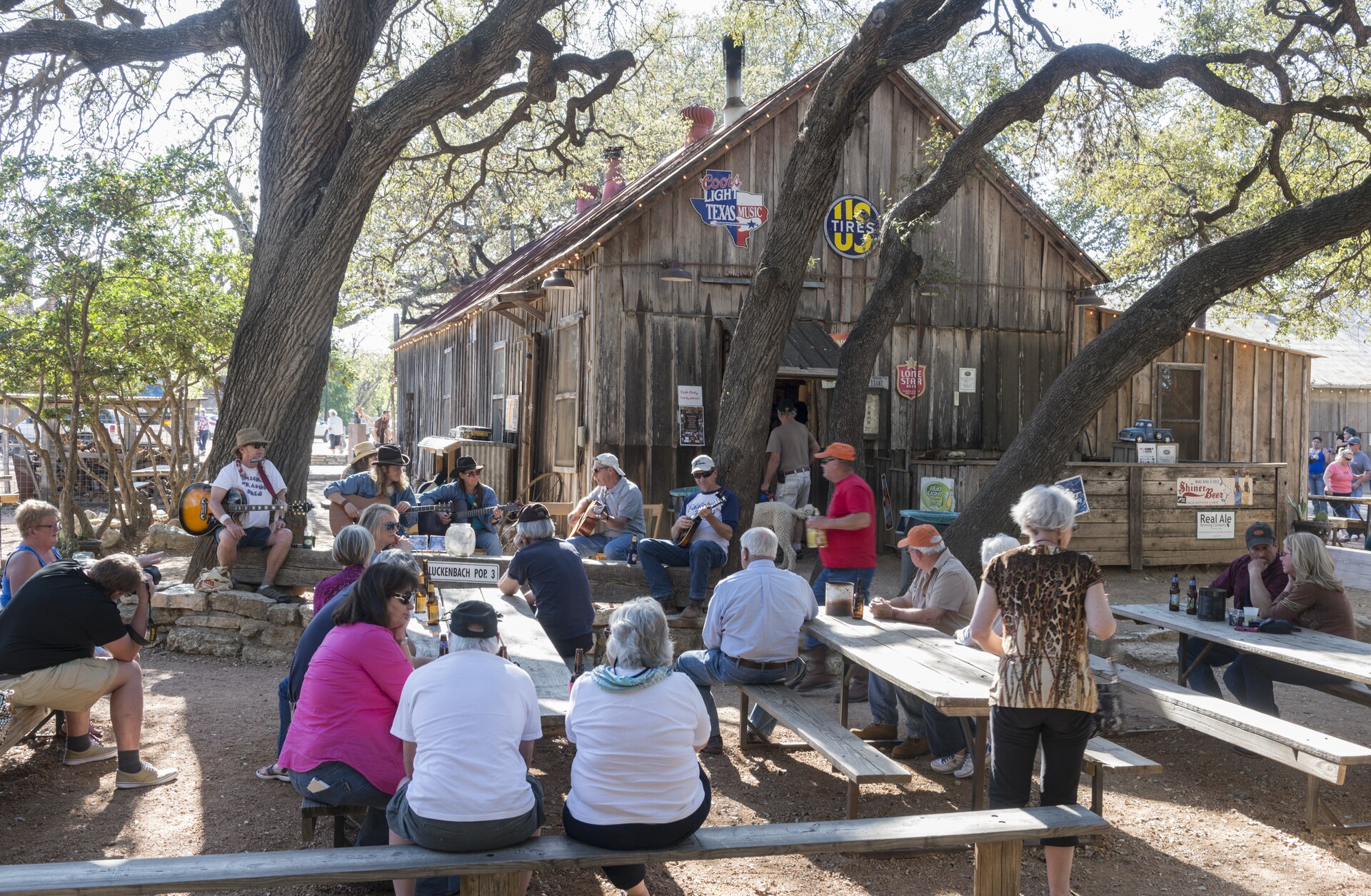 Visitors gather beneath live oaks for music, drinks, and barbecue outside Luckenbach's rustic general store in the Texas Hill Country
