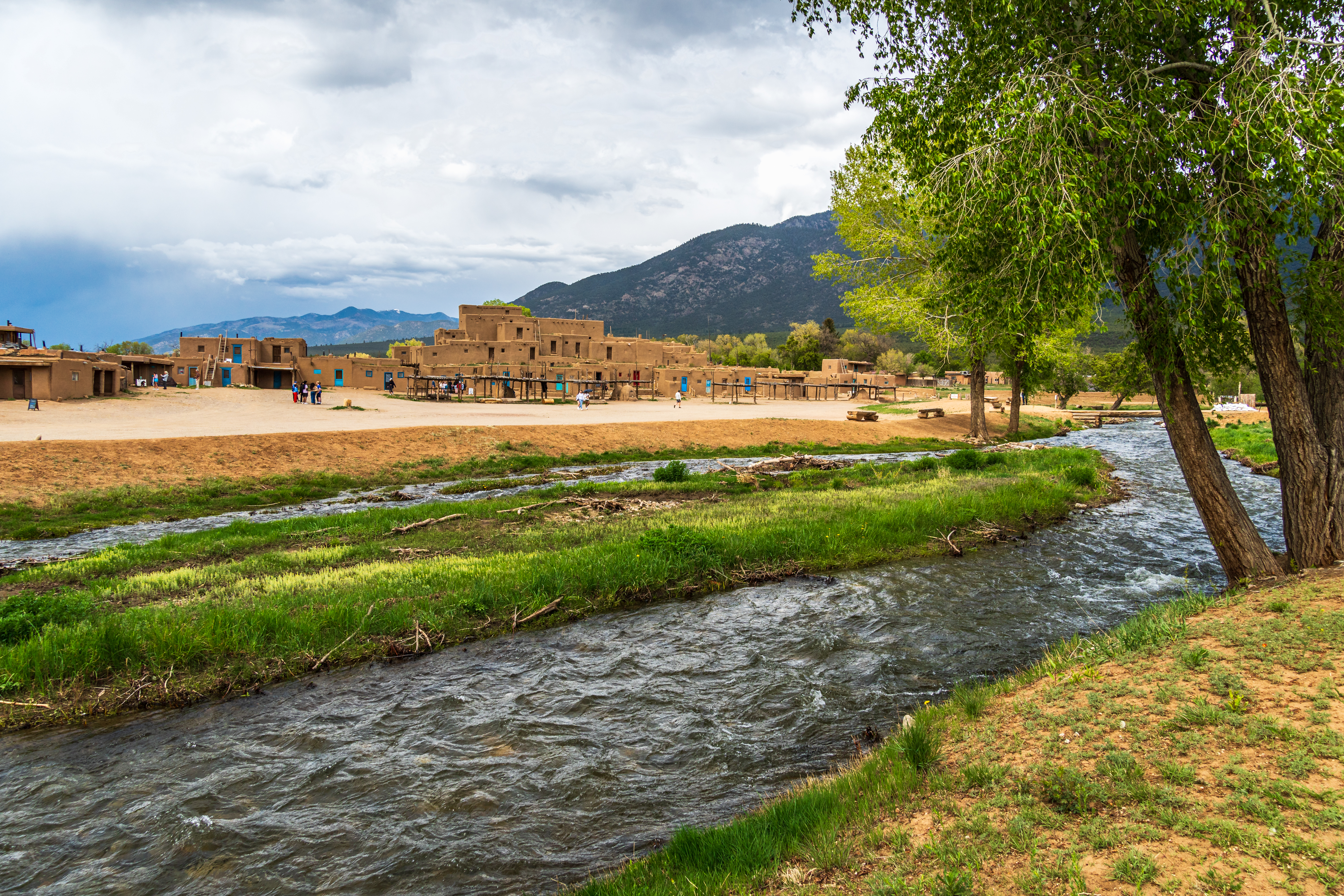 Taos Pueblo adobe buildings beneath the Sangre de Cristo Mountains beside flowing water