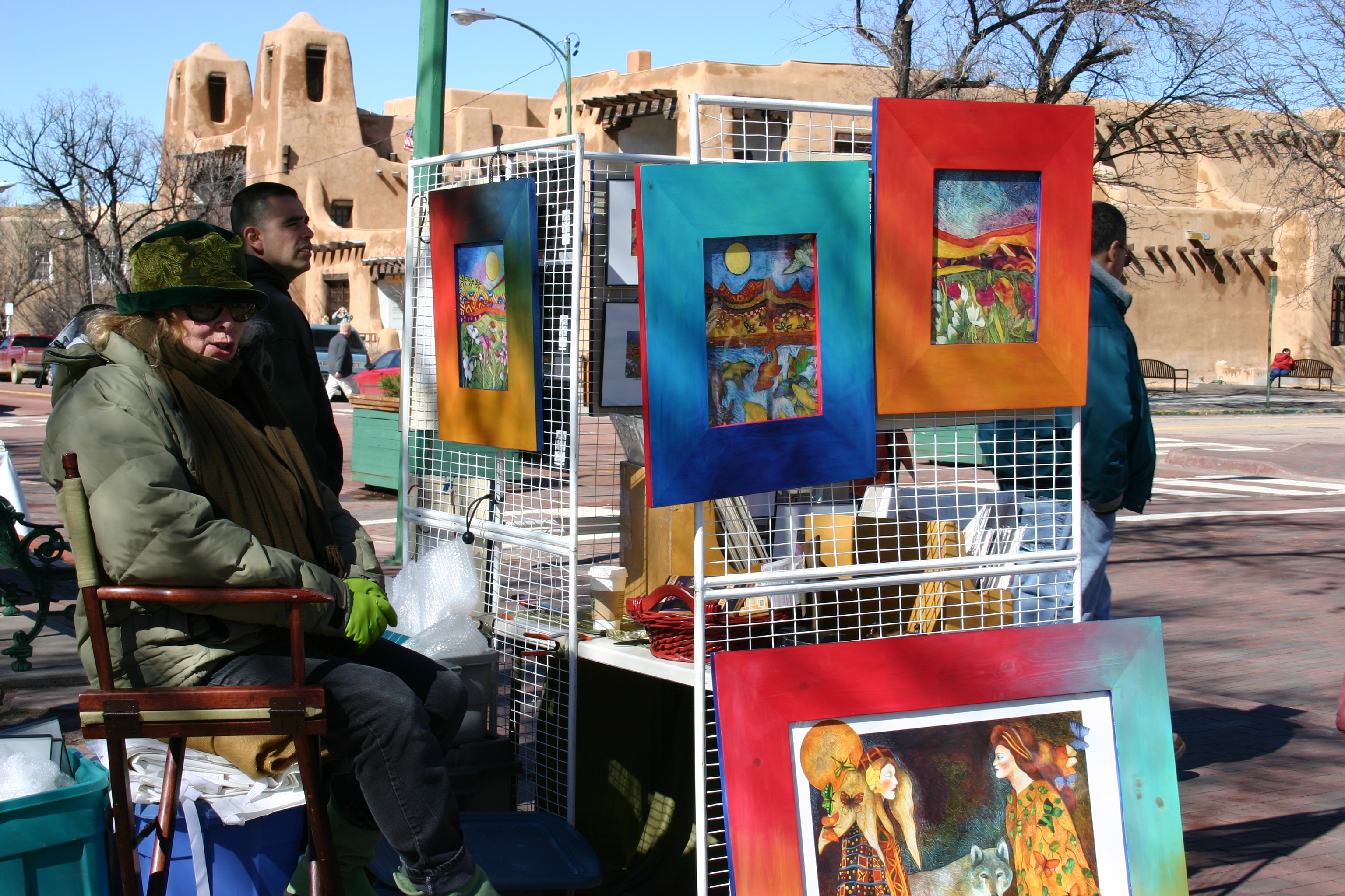 A Santa Fe Plaza artist displaying colorful work beside visitors and Pueblo Revival adobe architecture